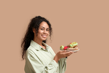 African-American woman with tasty eclairs on beige background