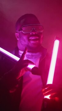 Vertical video. Rap show. Nightclub performance. Cool stylish confident joyful man in trendy sunglasses singing at hip hop event stage on bright neon lamp light smoke background.