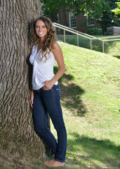 Stunning young Hispanic woman in white tank top and blue jeans - casual fashion - posing in park next to tree