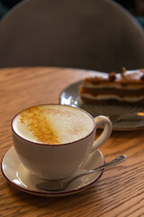 Side view of porcelain cup of pumpkin latte made from espresso, milk, ground pumpkin seeds and ground nutmeg standing on wooden table by chocolate cake. Soft focus. Copy space. Coffee break theme.