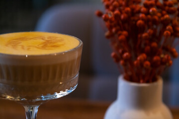 Close-up side view of glass dessert saucer full of pumpkin latte made from espresso, milk, pumpkin puree and ground dry mango standing on wooden table by vase with red decoration in coffeehouse.
