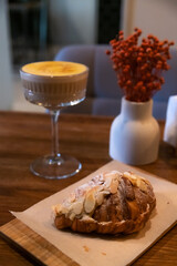 Close-up view of pumpkin latte with mango in glass dessert saucer, almond croissant and white vase with red decoration standing on wooden table in cafe or restaurant. Soft focus. Autumn food and drink