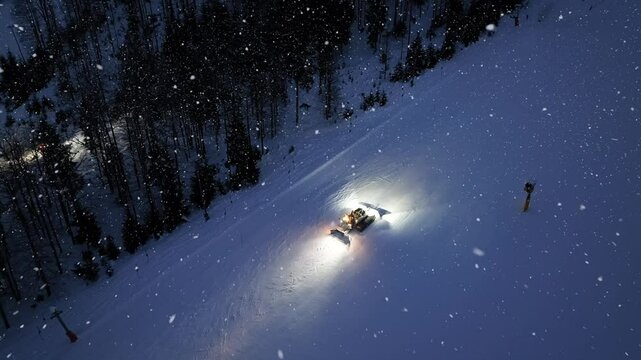 Aerial view of working snowcat on snowy alpine ski slope at winter night in snowfall. Kranjska Gora, Slovenia. Top drone view of snowcat grooming snow, ski resort, city lights at dusk. Snowplough