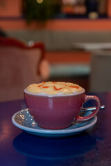 Close-up view of purple porcelain cup of pumpkin latte made from espresso, milk, whipped cream and caramel sprinkles standing on blue table in cafe or restaurant. Inspirational coffee drinks theme.
