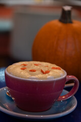 Close-up view of purple porcelain cup of pumpkin latte made from espresso, milk, whipped cream and caramel sprinkles standing on blue table next to orange pumpkin fruit in cafe or restaurant.