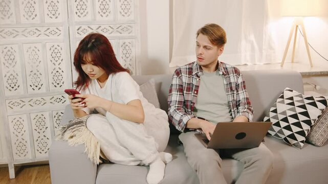 Husband and asian wife arguing on sofa, a man accusing his girlfriend of adultery, showing a smartphone screen with messages from her lover