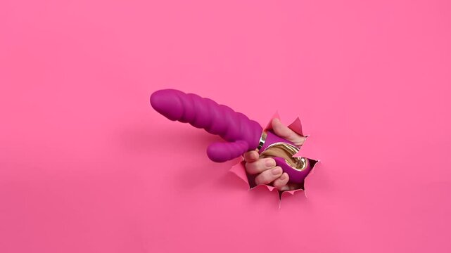 Female hand sticking out with pink sex toy from pink paper background. Copy space. 