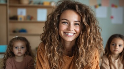 A radiant woman with long curly hair joyfully smiles, exuding warmth and kindness, sitting in a classroom with children in the backdrop filled with books.