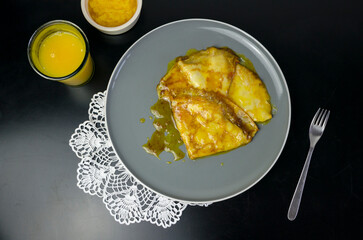 Crepes Suzette, a French dessert consisting of sauce of caramelized sugar and butter, orange juice and orange liqueur, on a black table, white crochet napkin, grey plate, top view