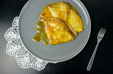 Crepes Suzette, a French dessert consisting of sauce of caramelized sugar and butter, orange juice and orange liqueur, on a black table, white crochet napkin, grey plate, top view