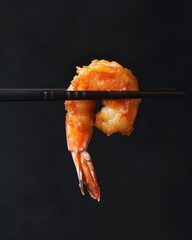 Close up of pair of chopsticks holding fried fresh tempura shrimp or prawn isolated on solid black background, healthy asian seafood cuisine.