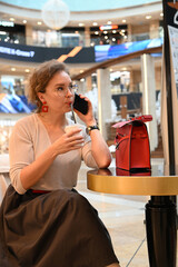 A young woman of European appearance is talking on the phone over a glass of coffee at a table in a shopping center cafe