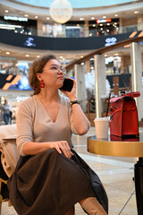 A young woman of European appearance is talking on the phone over a glass of coffee at a table in a shopping center cafe