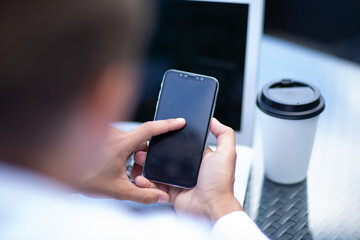 A man is holding a cell phone and looking at it. A cup of coffee is on a table next to him