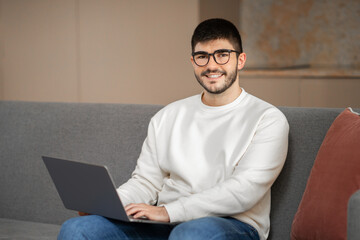 A young Caucasian man, wearing eyeglasses, smiles while sitting on a gray couch. He is focused on his laptop, immersed in a comfortable home environment.
