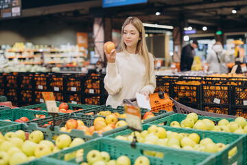Woman choosing apples in supermarket holding shopping list