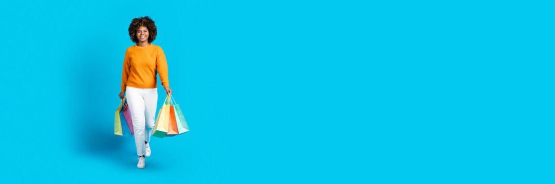 Sale season. Happy cheerful beautiful young black woman in stylish outfit shopaholic walk with purchases colorful shopping bags over blue studio background, smiling at camera, copy space, full length