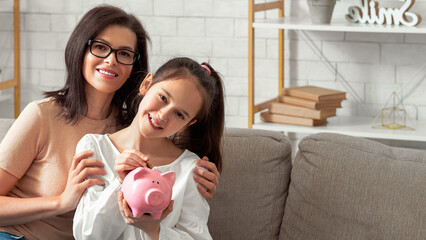 Happy young mother and her teen daughter holding piggybank, sitting on sofa at home, copy space....