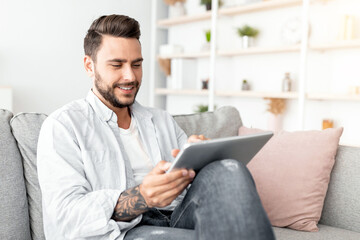 Relaxed millennial man resting on sofa with digital tablet, free space. Positive guy browsing social networks or shopping online while sitting in living room interior