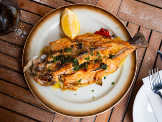 On wooden table there is plate with dinner of sea fish - baked trout carcass, complemented with asparagus, baked potatoes, slice of lemon.