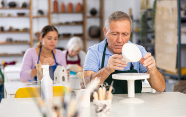 elderly man potter carefully paints vase in white