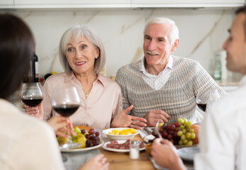 Mature spouses sitting at table, enjoying food and wine, chatting talking laughing with adult son and his girlfriend. In homely atmosphere male and female pensioner celebrate anniversary with guests
