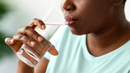 Cropped view of overweight black female drinking mineral water from glass indoors, panorama. Plus...