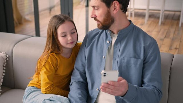 Happy family smiling Caucasian man guy dad talk to little girl at home daddy male parent teaching daughter kid use mobile phone device father child ordering food online browsing network social media