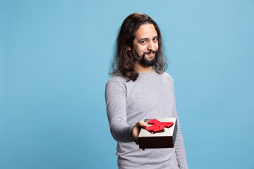 Sweet affectionate guy offering a gift in a cute box with ribbon on camera, giving presents for Christmas or anniversary against blue background. Person in casual wear showing gratitude and love.