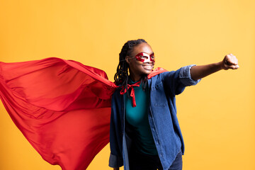 African american girl wearing superhero costume and mask for Halloween doing flying pose, studio background. Woman dressed as comic book hero hiding identity doing heroic hand gesture © DC Studio