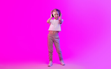 A preteen girl stands confidently with headphones on, striking a stylish pose against a vibrant pink background. She embodies a relaxed, fun lifestyle while enjoying her music.