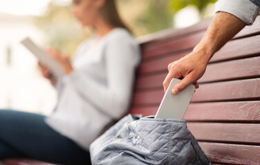 Pickpocket Thief Stealing Mobile Phone From Woman's Bag While She Sitting On Bench In Park. Cropped, Selective Focus