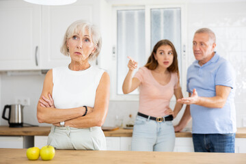 Fototapeta premium Elderly frustrated woman turned away from scolding her husband and adult daughter in the kitchen