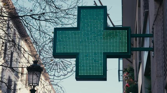 A vibrant green LED pharmacy cross sign stands out on a street corner in Madrid, Spain. The bright light indicates the presence of a nearby pharmacy, a common feature in European cities, symbolizing