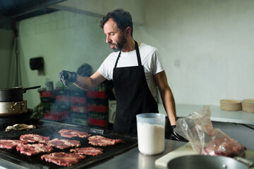 Latin chef sprinkling salt on meat cooking on a grill in a commercial kitchen, preparing beef tacos
