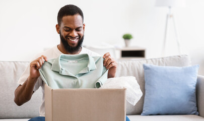 Shopping Delivery. Joyful African American Man Receiving Clothes Unpacking Cardboard Box And Holding New T-Shirt Sitting On Couch At Home. Happy Customer Uboxing Delivered Purchase. E-Commerce