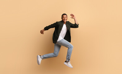A joyful African American man is leaping in a dynamic pose. He wears casual attire with jeans and a shirt, showcasing an upbeat lifestyle while posed against a soft, beige backdrop.