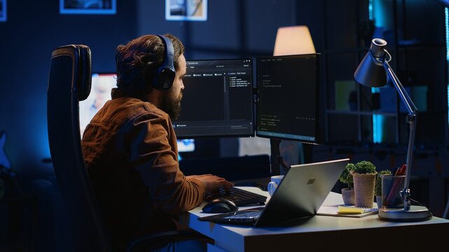 Engineer typing code in apartment office, using javascript programming language to develop software application. It admin using multimonitor desktop setup and notebook to finish tasks, camera A