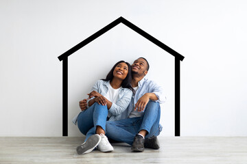 Planning relocation. Dreaming african american couple sitting near white wall with drawn house and...