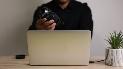 Photographer holding camera lens while working on laptop in modern office