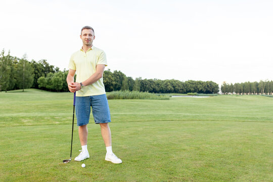 young sporty man stands on golf course holding club and looking at the camera, guy golfer plays golf on green lawn