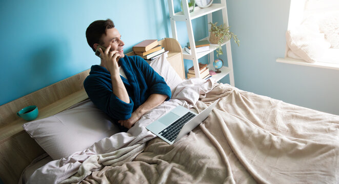 A man in a blue robe sits on a bed, talking on the phone with a laptop nearby. A cozy bedroom with books, a plant, and natural light from a window creates a relaxed, productive vibe.