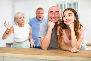 Portrait of a happy married couple in a home kitchen, not paying attention to adult family members...