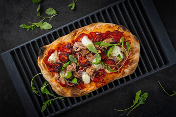 Traditional Italian pinsa bread prosciutto e salsa di pomodoro with tuna fish, buffalo mozzarella and onion rings served with tomato sauce as top view on an old rustic grill grid