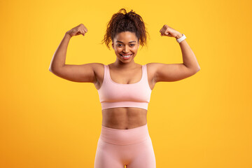 A young woman stands confidently with her arms flexed. She wears a pink workout set that highlights her fitness. The bright yellow background enhances her vibrant energy and strength.