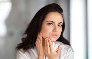 A young brunette woman examines her skin in a stylish bathroom, focusing on her skincare routine. She is engaged in self-care, reflecting a commitment to beauty and wellness.