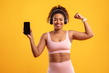 A young woman is smiling confidently while wearing workout attire and headphones. She holds a smartphone in one hand and flexes her bicep with the other, showcasing her fitness enthusiasm.