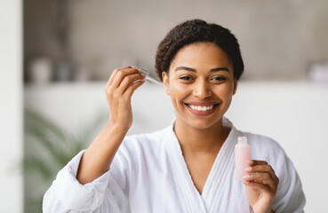 African American woman in a white bathrobe smiles as she applies serum to her face using a dropper. The modern bathroom is bright and airy, enhancing the relaxing atmosphere of her skincare routine.