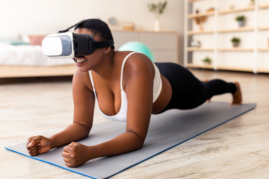 Domestic virtual training. Curvy young black lady in VR glasses standing elbow plank at home. Overweight millennial woman training via augmented reality app. Sports and technologies concept