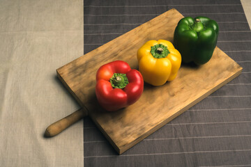 Raw yellow, red and green peppers on wooden cutting board above the table